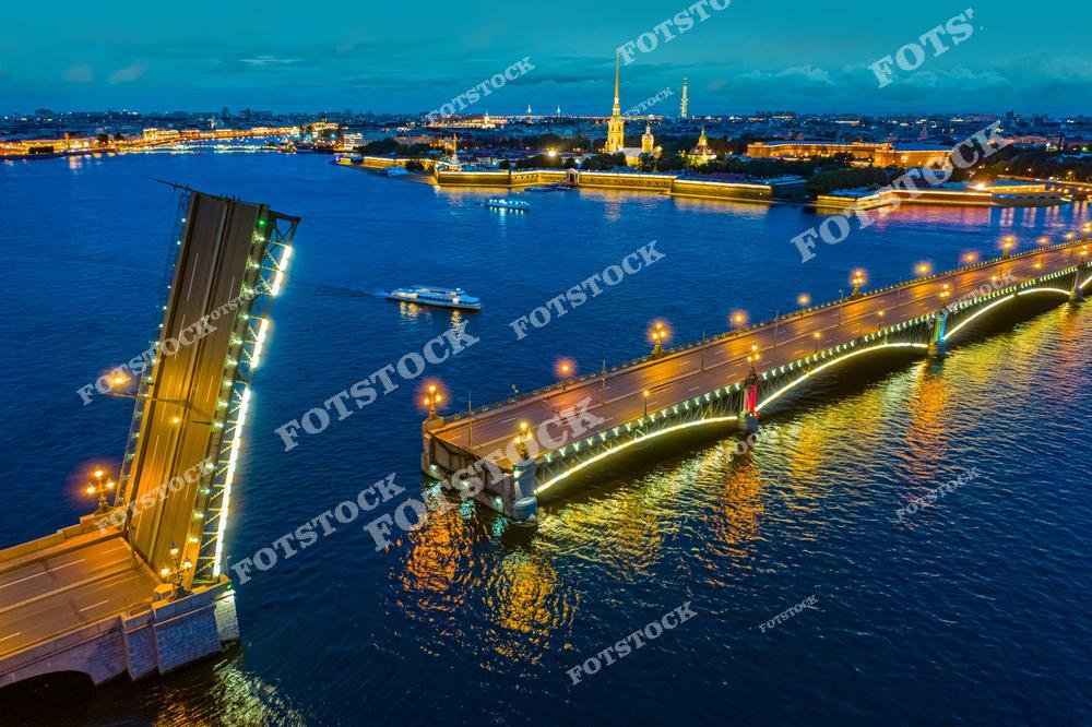 На фотографии запечатлён Троицкий мост в момент разведения, один из красивейших мостов Санкт-Петербурга. Подсветка пролётов отражается в водах Невы, создавая эффектный световой рисунок. На заднем плане видна Петропавловская крепость с её шпилем и вечерним освещением. По реке движется прогулочное судно. Снимок сделан с воздуха, подчёркивая масштаб и атмосферу ночного города.